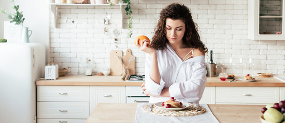 panoramic shot of girl holding orange and looking at pancakes in kitchen