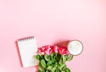 The cup of hot coffee, empty notepad with funny pen and bouquet of fresh roses on soft pink background.