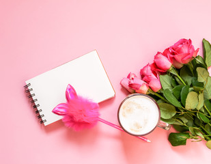 The cup of hot coffee, empty notepad with funny pen and bouquet of fresh roses on soft pink background.