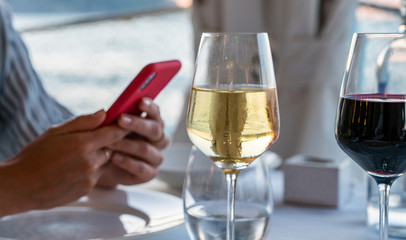 Woman's hands holding smartphone during dinner. Beautifully served table with wine glass on the Mediterranean restaurant table. Holidays dinner outdoors overlooking the sunset.