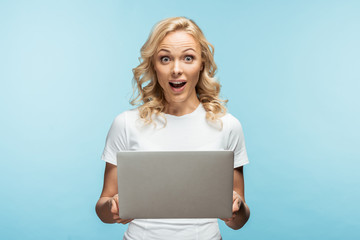 Naklejka premium surprised blonde woman holding laptop and looking at camera on blue