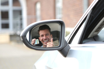 Junger Mann schaut lachend in den Aussenspiegel seines Autos und zeigt einen Führerschein