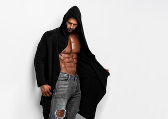 Handsome macho man posing shirtless in unbuttoned coat with hood exposing his muscular build, in...