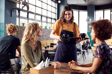 Two Female Friends Sitting At Table In Coffee Shop Being Served By Waitress