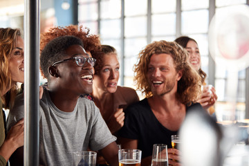 Group Of Male And Female Friends Celebrating Whilst Watching Game On Screen In Sports Bar