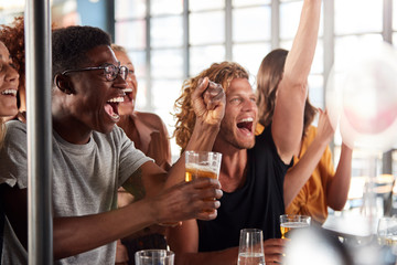 Group Of Male And Female Friends Celebrating Whilst Watching Game On Screen In Sports Bar