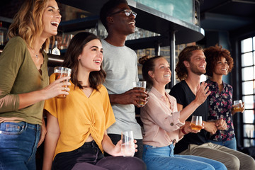 Group Of Male And Female Friends Celebrating Whilst Watching Game On Screen In Sports Bar