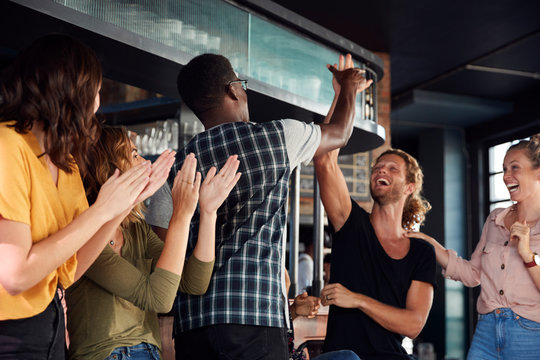 Group Of Male And Female Friends Celebrating Whilst Watching Game On Screen In Sports Bar - Powered by Adobe