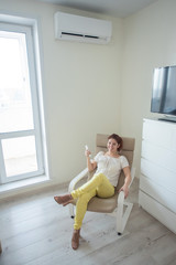 Girl in bright clothes and shoes of the color of a dusty rose sits in an armchair with a phone in her hands. A young woman sits in a bright room and is cooled on a hot day using an air conditioner.