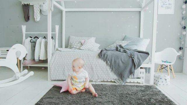 Joyful toddler girl sitting on carpet and trying by herself to get up from floor in kid's room. Adorable infant taking first steps learning to walk, firstly trying to stand without support at home.