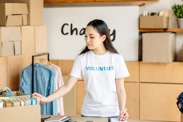 attentive asian volunteer writing in notebook while standing near rack with shirts