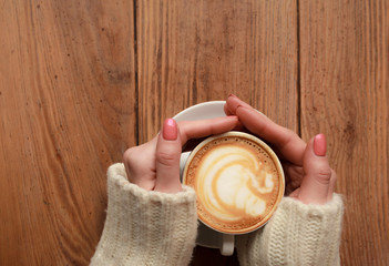 Lonely woman with nice manicure in warm wood sweater drinking coffee in the morning, top view of female hands holding cup of hot beverage on wooden desk, retro toned.