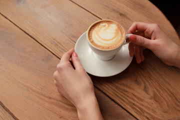 Lonely woman with nice manicure in warm wood sweater drinking coffee in the morning, top view of female hands holding cup of hot beverage on wooden desk, retro toned.