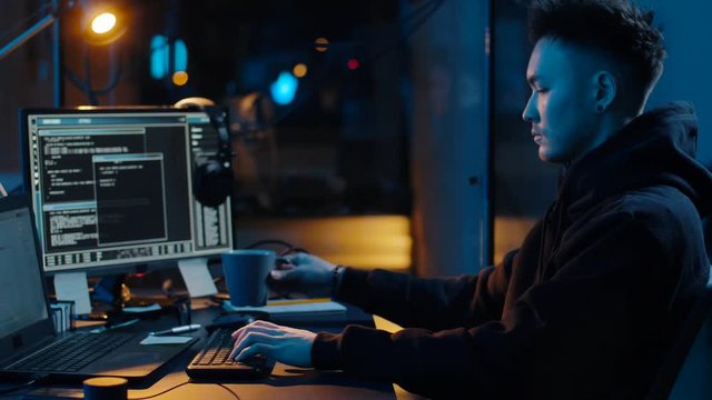 cybercrime, hacking and technology concept - asian male hacker writing code or using computer virus program for cyber attack and drinking coffee in dark room at night