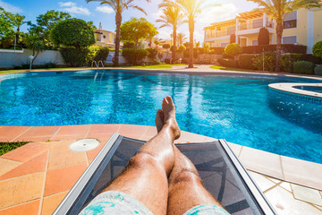 Relaxing at the swimming pool. Man relaxing next to swimming pool. Man enjoying the hot summer at swimming-pool. Sunbathing by the swimming pool, mans legs lying down on a sun lounger over the water. - obrazy, fototapety, plakaty