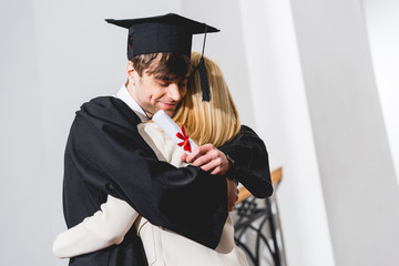 cheerful son in graduation cap holding diploma while hugging blonde mother