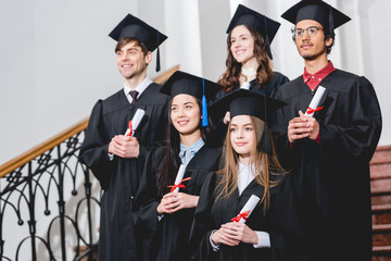 cheerful students in graduation gowns holding diplomas in university