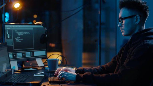 cybercrime, hacking and technology concept - asian male hacker in glasses writing code or using computer virus program for cyber attack and drinking coffee in dark room at night