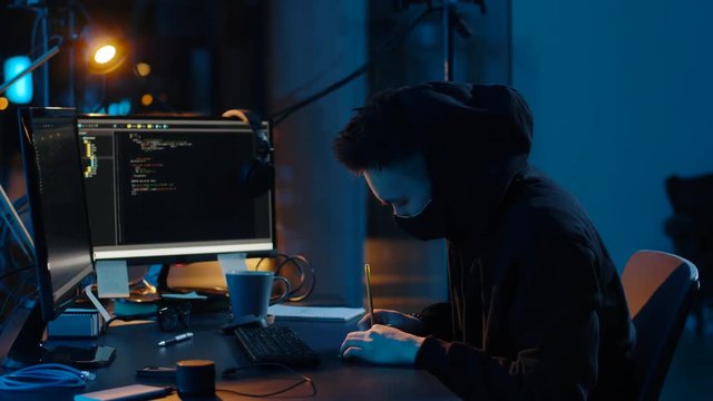 cybercrime, hacking and technology concept - asian male hacker in mask using computer virus program for cyber attack and taking notes in dark room at night