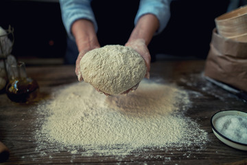 Female hands holding dough in ball shape. Basic homemade dough with ingredients on the side on wooden table with natural light. Home healthy food. Selective focus. Focus on the dough.