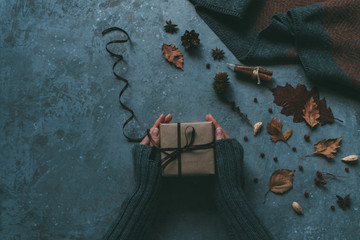 Autumn background. Handmade gift box in a woman's hand wrapped a craft paper on the grey background with falling leaves, cinnamon sticks and cones. Hello autumn. Instagram fashion composition.