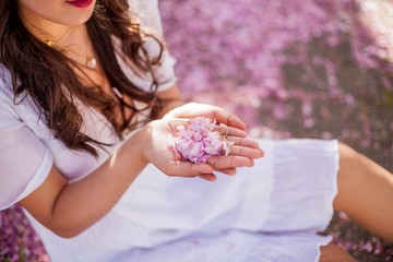 Close-up. Female hands hold petals of pink flowers. A young beautiful Asian woman in a white dress walks in a flowered park.
