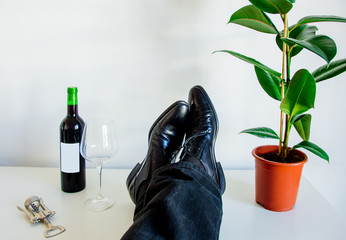 Male foots in low shoes and ficus plant with next to glass and red wine bottle on white table in a...