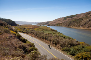 Two cross country bike riders ride along road parallel to a river in California. 