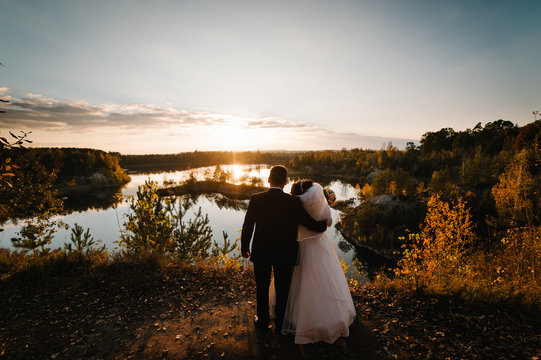 The wedding couple is standing back by the lake and looking at the landscape. Autumn nature and sunset. The groom embraces the bride hug.