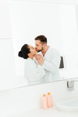 happy woman kissing with boyfriend while standing near mirror in bathroom