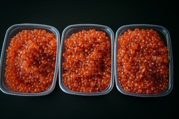 Red caviar in plastic containers close up, top view, black background. Gourmet food.