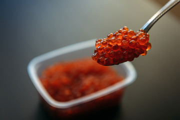 Red Caviar in a spoon and in plastic container over dark background. Close-up salmon caviar. Delicatessen. Seafood.