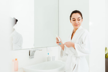 selective focus of brunette woman standing near mirror while holding toothbrush and toothpaste