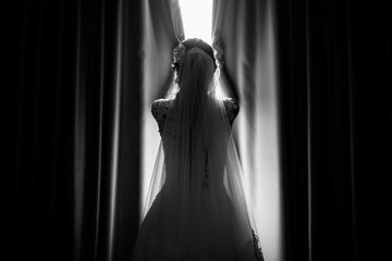 The bride touches the curtains while standing near the palace window. beautiful bride in white dresses opening curtains. Rear view. Black and white photo. Lens blur, film noise