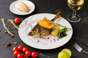 stuffed fish with carrots, parsley, horseradish and salad on a white plate, with tomatoes, lime, fork and knife, with a glass of white wine on a black background
