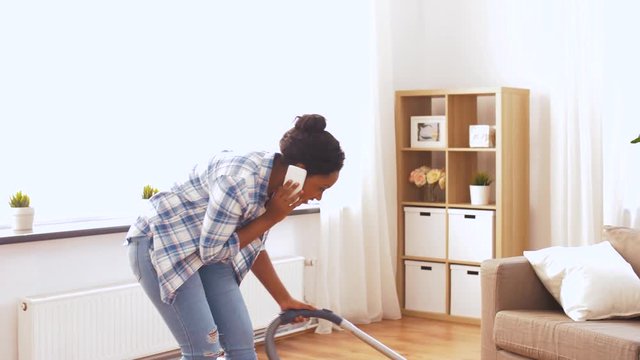 people, housework and housekeeping concept - happy african american woman with vacuum cleaner calling on smartphone at home