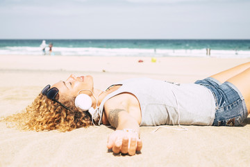 Happy people cheerful blonde beautiful curly woman lay down and relax enjoying the beach in summer holiday vacation taking a sunbath and listenint music with headphones - ocean and sky in background