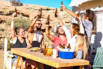 Friends people toasting together with fun in outdoor alternative rural camping site - cheerful young men and women laughing and smiling under the sun with beer bottles - country side place