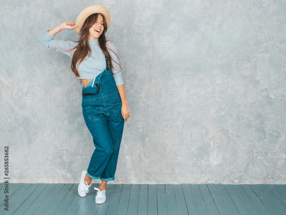 Wall mural Portrait of young beautiful smiling woman looking at camera. Trendy girl in casual summer overalls clothes and hat. Funny and positive female posing near gray wall in studio. Full length