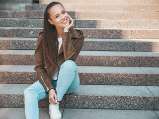 Portrait of beautiful smiling brunette model dressed in summer hipster jacket and jeans clothes. Trendy girl sitting on steps in the street background. Funny and positive woman in round sunglasses