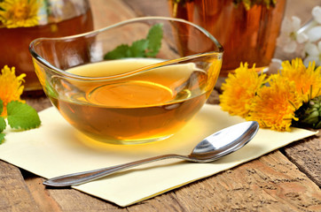 Close-up of dandelion honey in a glass bowl and dandelion head around, jar and herbal tea in background