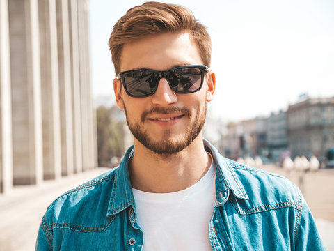 Portrait of handsome smiling stylish hipster lumbersexual businessman model. Man dressed in jeans jacket clothes. Fashion male posing on the street background in sunglasses
