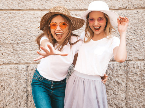 Portrait of two young beautiful blond smiling hipster girls in trendy summer white t-shirt clothes. Sexy carefree women posing in the street near wall. Positive models having fun in sunglasses and hat