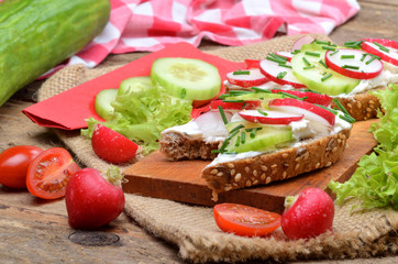 Wholemeal grain bread with curd cheese, fresh radish, cucumber and tomatoes on a wooden cutting board - concept of healthy fitness breakfast or snack, salad and whole cucumber in background
