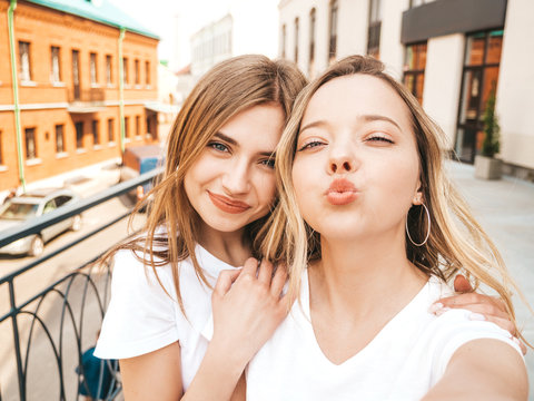 Two young smiling hipster blond women in summer white t-shirt clothes. Girls taking selfie self portrait photos on smartphone.Models posing on street background.Female making duck face