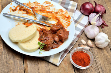 Eating pork goulash meat with dumplings on white plate, cutlery, garlic, onion, pepper, tablecloth in the background - typical Czech food