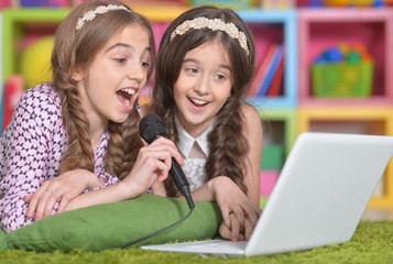 Portrait of two cute little girls singing karaoke with laptop while lying on floor