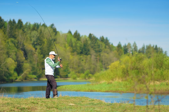 Fisherman with a spinning rod catching fish on a river at sunny summer day with green trees at background. Outdoor weekend activity. Photo with shallow depth of field taken at wide open aperture.