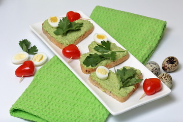 Sandwiches for breakfast. Heart-shaped bread slices smeared with ground avocado. Decorated with boiled quail eggs, parsley and tomatoes. Unfolded on a white rectangular plate.