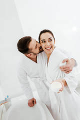 Fototapeta premium cheerful man hugging and kissing cheek of happy girlfriend holding container with face cream